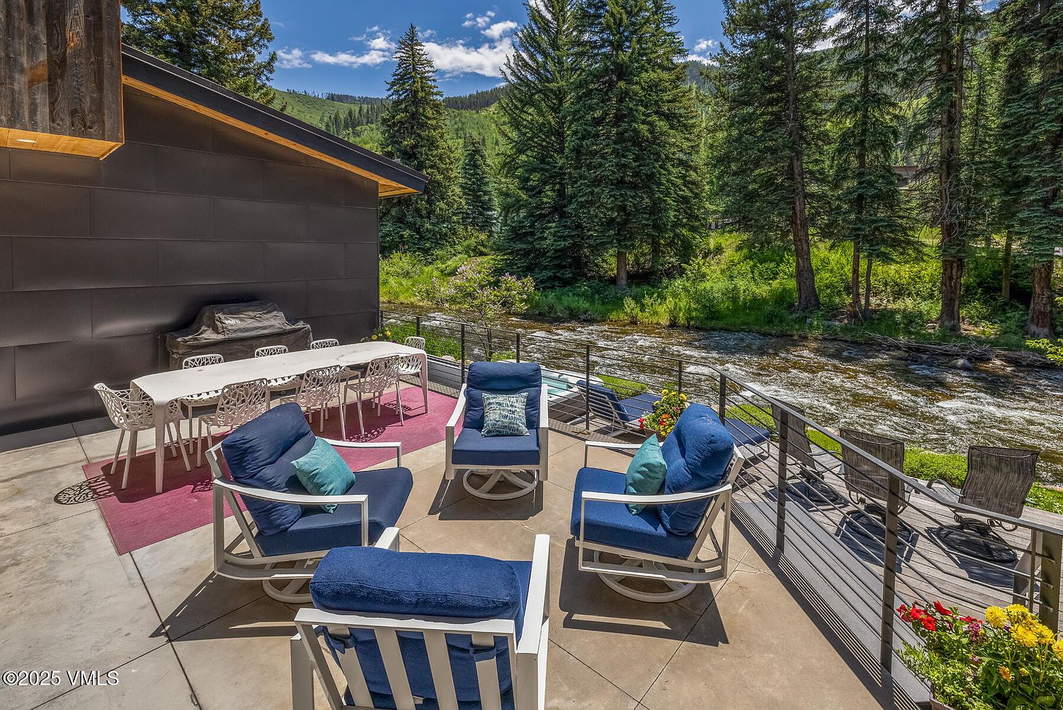 This expansive outdoor patio features a modern dining set and a comfortable seating area with blue-cushioned chairs, all set against a backdrop of a rushing mountain stream. The space is framed by a sleek metal railing and overlooks a lush, tree-lined landscape, creating a serene and private atmosphere. The contemporary architecture of the home, characterized by dark paneling, complements the natural beauty of the surrounding forest.