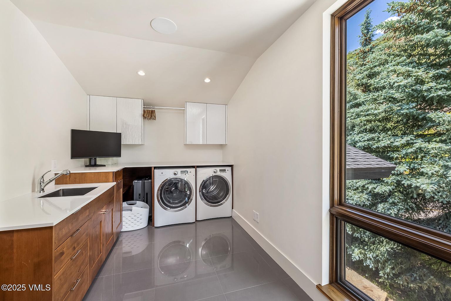 This modern laundry room features sleek, dark-toned tile flooring and warm-toned wood cabinetry topped with a clean, light-colored countertop. The space includes a built-in sink, a side-by-side washer and dryer set, and wall-mounted cabinets for storage. A large window on the right provides ample natural light and a view of the surrounding trees, creating a bright and functional workspace.