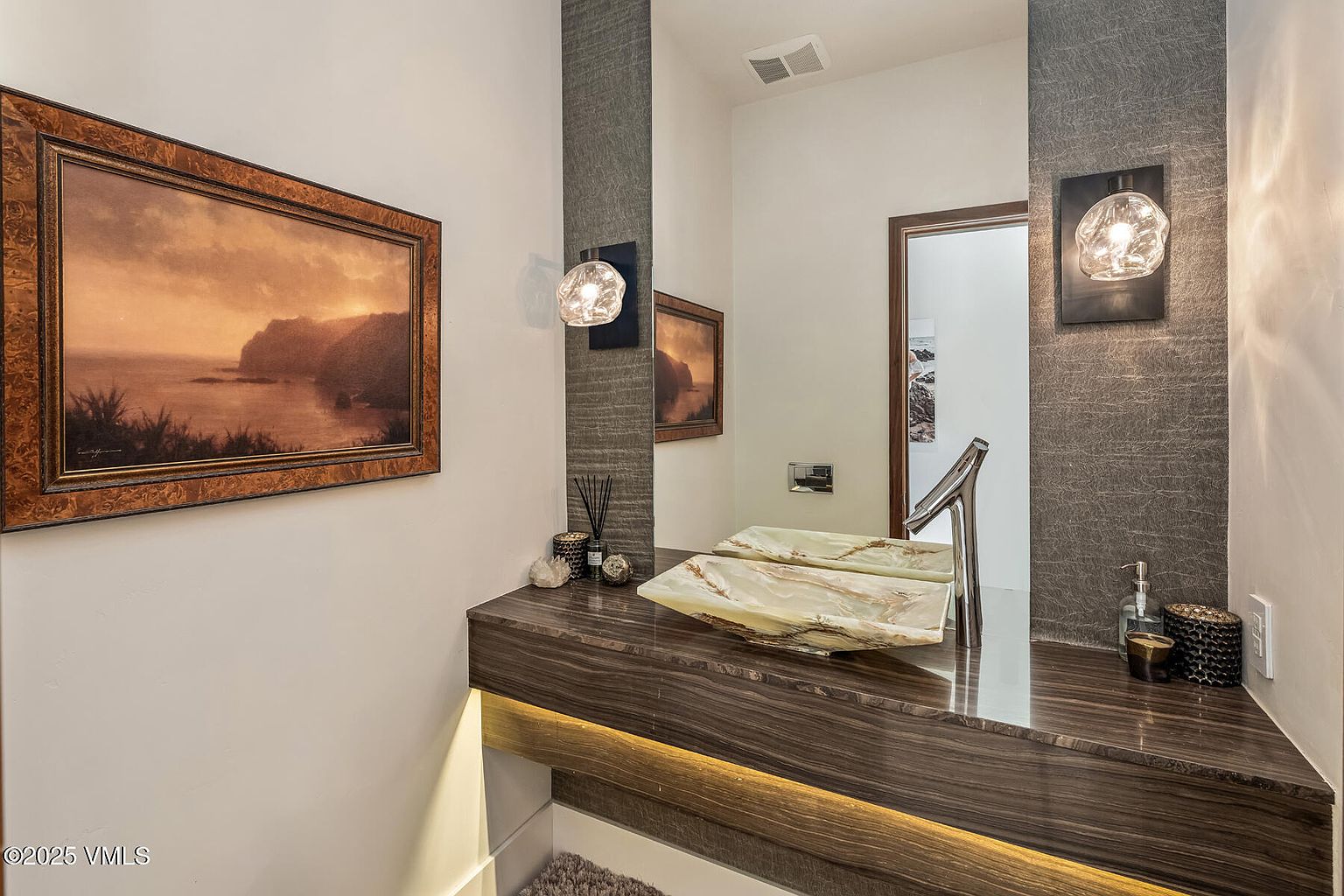 This elegant powder room features a floating vanity with a dark wood-toned finish and a unique, translucent stone vessel sink. A large, frameless mirror is mounted against a textured, dark gray accent wall, flanked by two modern, orb-style wall sconces. The space is illuminated by warm under-cabinet lighting, creating a sophisticated and moody atmosphere.