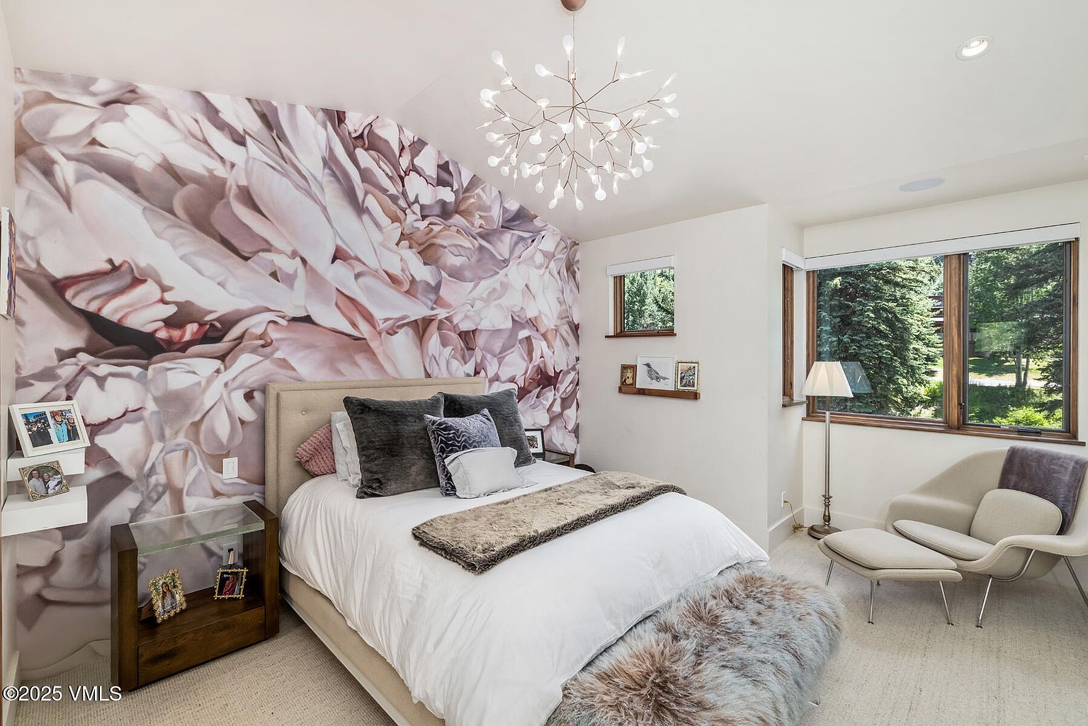 This cozy bedroom features a striking floral-patterned accent wall behind a neutral upholstered bed frame. The room is illuminated by a modern, branching chandelier and includes a comfortable armchair with an ottoman near a large window overlooking trees. A plush, textured bench sits at the foot of the bed, adding a layer of warmth and luxury to the space.