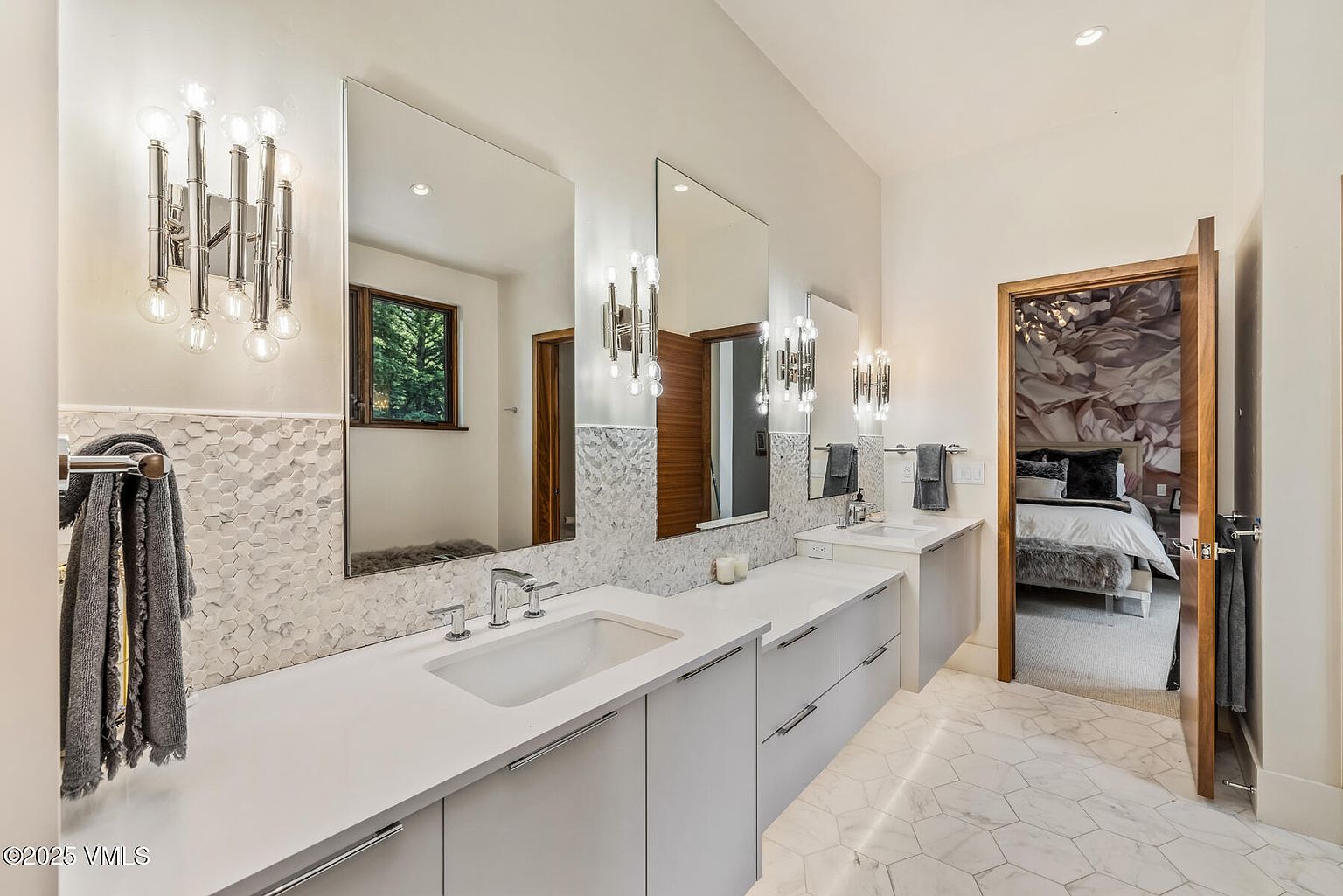 This modern primary bathroom features a long, light-toned vanity with dual sinks and sleek, contemporary light fixtures mounted on mirrors. The walls are adorned with a textured, geometric tile backsplash, while the floor is covered in large-format, light-colored patterned tile. A doorway leads into an adjacent bedroom, providing a glimpse of a decorative floral wall mural and a plush bed.