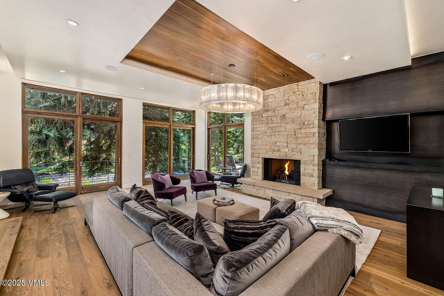 This spacious living room features a large, plush sectional sofa facing a stone-clad chimney breast with a built-in fireplace. A striking circular chandelier hangs from a recessed wood-paneled ceiling section, while tall windows provide expansive views of the surrounding trees. The room is accented by two deep purple armchairs and a modern lounge chair, creating a sophisticated and comfortable atmosphere.