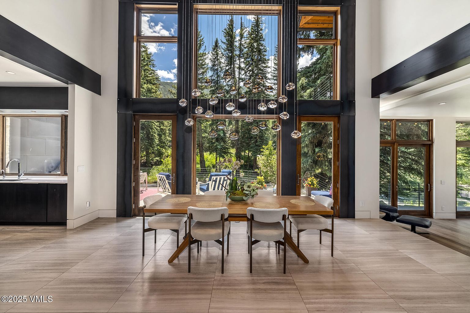 This elegant dining room features a large wooden table surrounded by four modern, light-toned chairs, positioned beneath a striking multi-pendant chandelier. The space is defined by expansive floor-to-ceiling windows that frame a lush forest view, seamlessly connecting the interior with the outdoor patio. The room is finished with neutral, large-format tile flooring and clean, minimalist walls, creating a bright and sophisticated atmosphere.