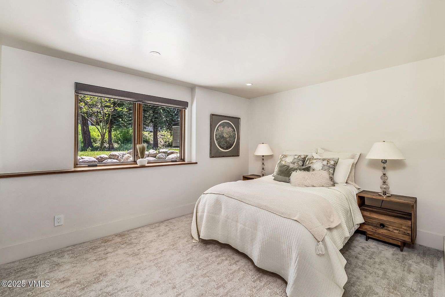 This cozy bedroom features a neutral color palette with a plush white bedspread and decorative pillows. A large window with a dark shade allows natural light to illuminate the room, offering a view of the outdoor greenery. The space is furnished with two rustic wood nightstands topped with matching table lamps, creating a warm and inviting atmosphere.