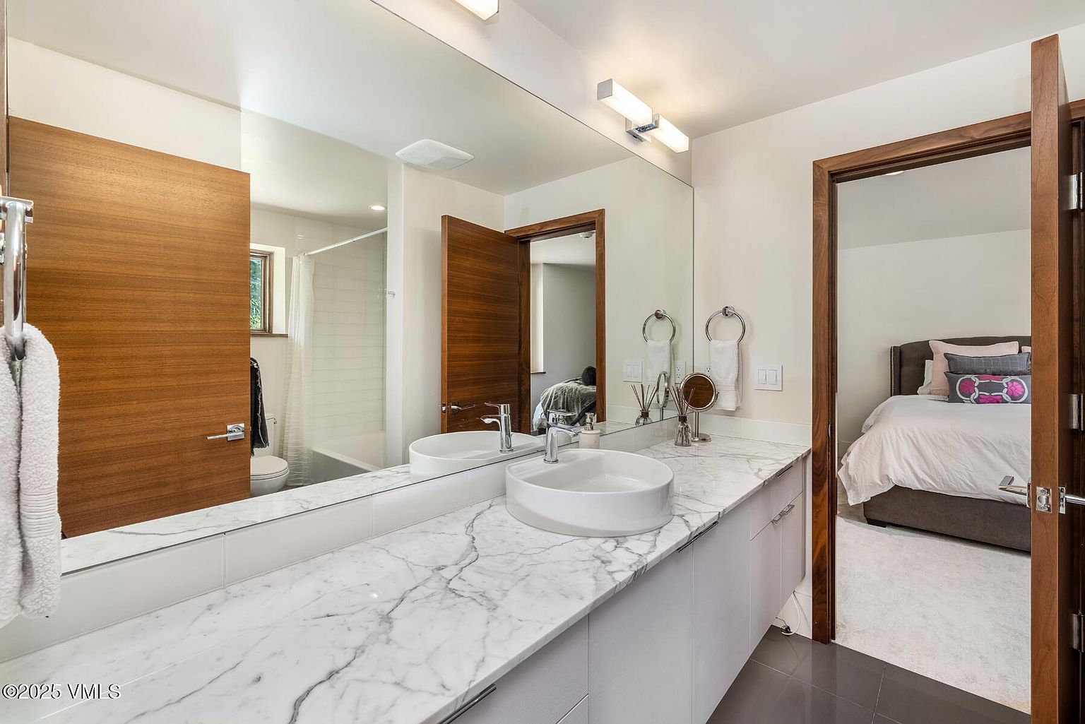 This modern bathroom features a sleek vanity with a white vessel sink set atop a patterned stone countertop. A large, expansive mirror reflects the space, including a glimpse into the adjacent bedroom, while warm wood-toned doors add a natural contrast to the clean, neutral walls. The perspective is from the doorway, capturing the vanity area and the transition into the bedroom suite.