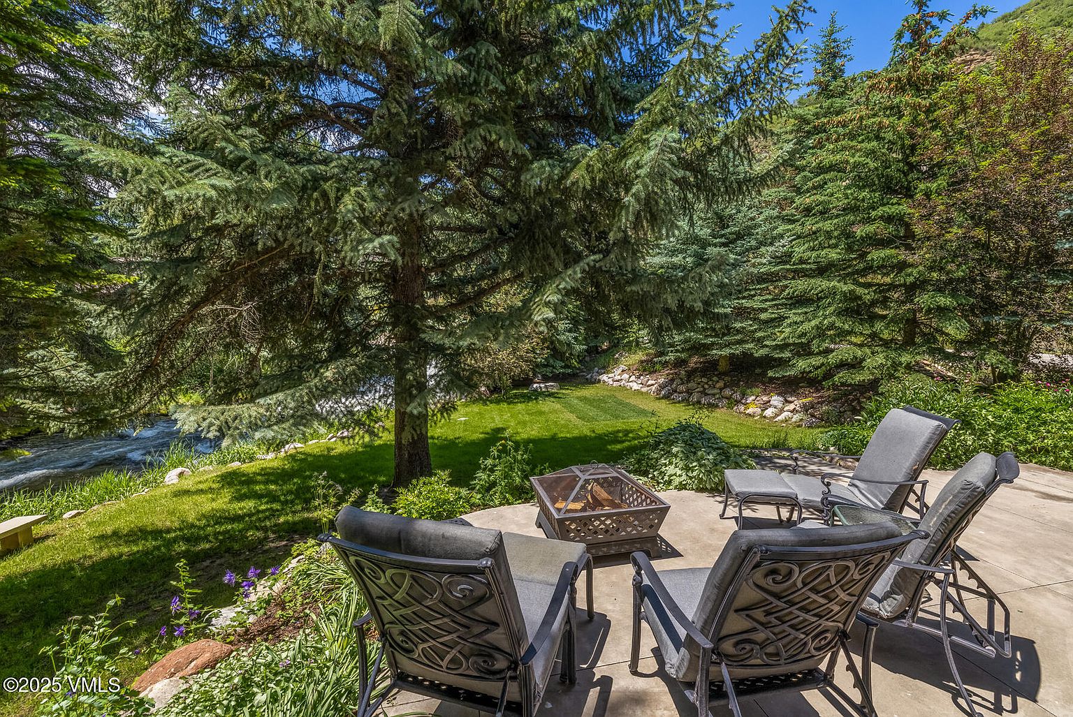 This serene outdoor patio space features a metal fire pit surrounded by comfortable lounge chairs, perfect for relaxing while overlooking a lush, green backyard. The area is shaded by mature evergreen trees and sits adjacent to a flowing stream, creating a peaceful and private atmosphere. The perspective captures the inviting seating arrangement in the foreground, leading the eye toward the natural landscape and water feature beyond.