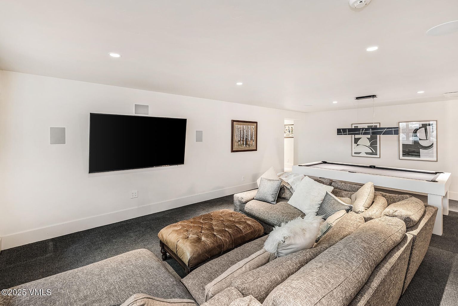 This spacious basement recreation room features a large, comfortable sectional sofa paired with a tufted leather ottoman, creating an inviting lounge area. A wall-mounted television serves as the focal point, while a white pool table sits in the background, adding a playful element to the space. The room is finished with neutral walls, recessed lighting, and dark carpeting, offering a versatile area for entertainment and relaxation.