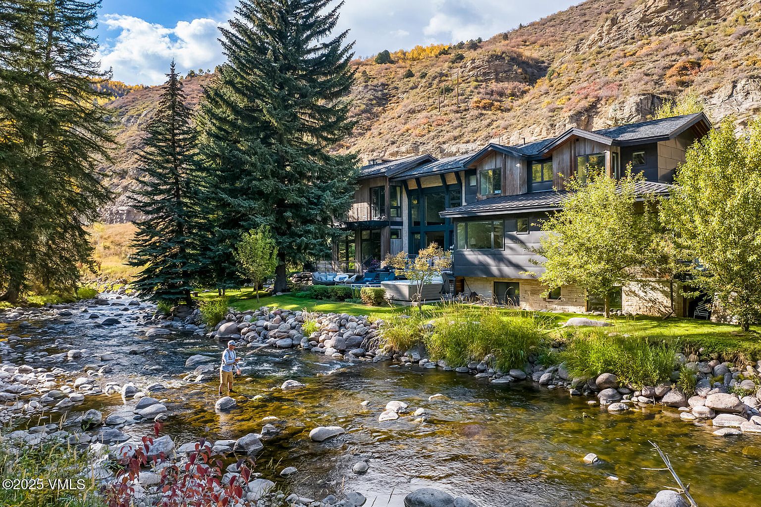This stunning mountain residence features a contemporary design with a mix of wood siding and stone accents, nestled along the banks of a clear, flowing stream. The rear of the home showcases large windows and multiple levels, with a landscaped lawn and a private hot tub area overlooking the water. A person is seen fly-fishing in the foreground, emphasizing the serene, outdoor-focused lifestyle offered by this property.