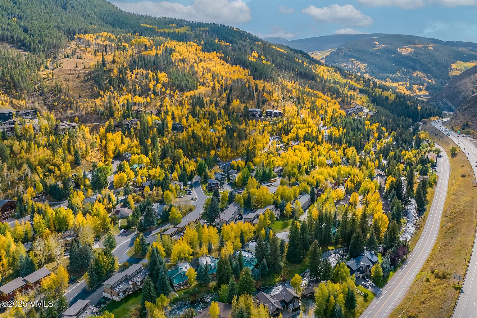 This high-angle aerial view captures a scenic mountain community nestled among vibrant autumn foliage. The landscape features a mix of residential homes and townhouses integrated into a forested hillside, with a winding road and a nearby highway running along the edge of the property. The scene is characterized by the striking contrast between the golden yellow aspen trees and the deep green pines, creating a picturesque and serene mountain setting.