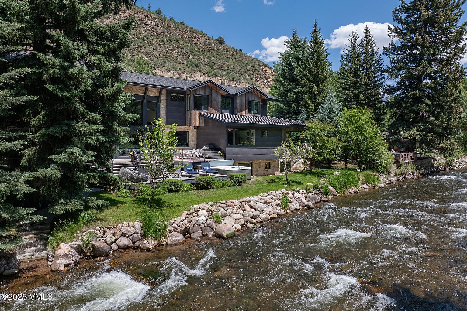 This rear view of a modern mountain residence showcases a multi-level architectural design with a blend of wood siding and stone accents. The property features a spacious deck with outdoor seating and a hot tub, overlooking a rushing river and a lush, tree-lined landscape. The setting is serene and private, emphasizing a seamless connection between the home's living spaces and the natural environment.
