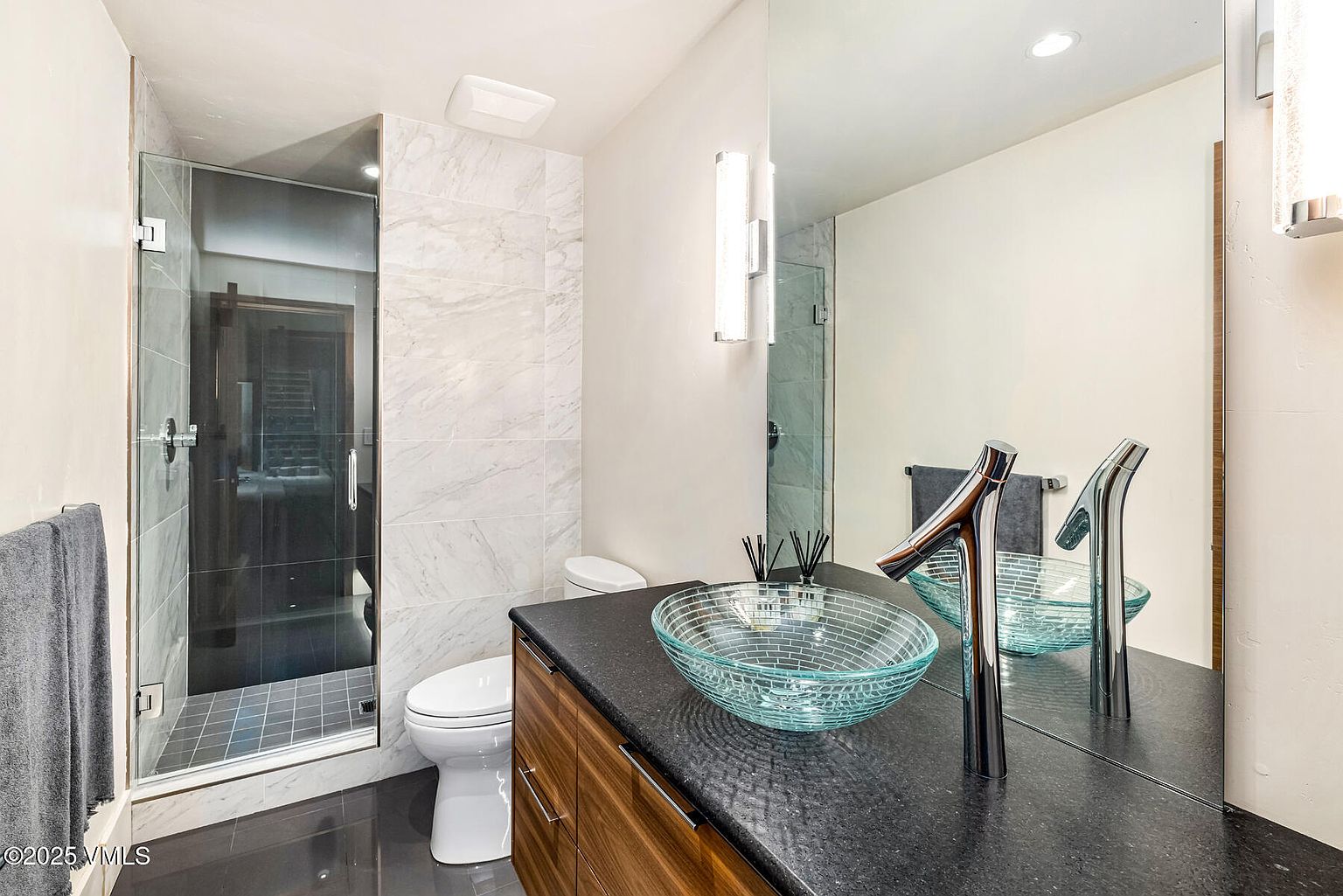 This contemporary bathroom features a striking glass vessel sink set atop a dark stone vanity with warm-toned wood cabinetry. A walk-in shower with a glass door is enclosed by light-colored stone wall tiles, while a large mirror reflects the vanity area to enhance the sense of space. The room is finished with dark floor tiles and modern chrome fixtures, creating a sleek and polished aesthetic.
