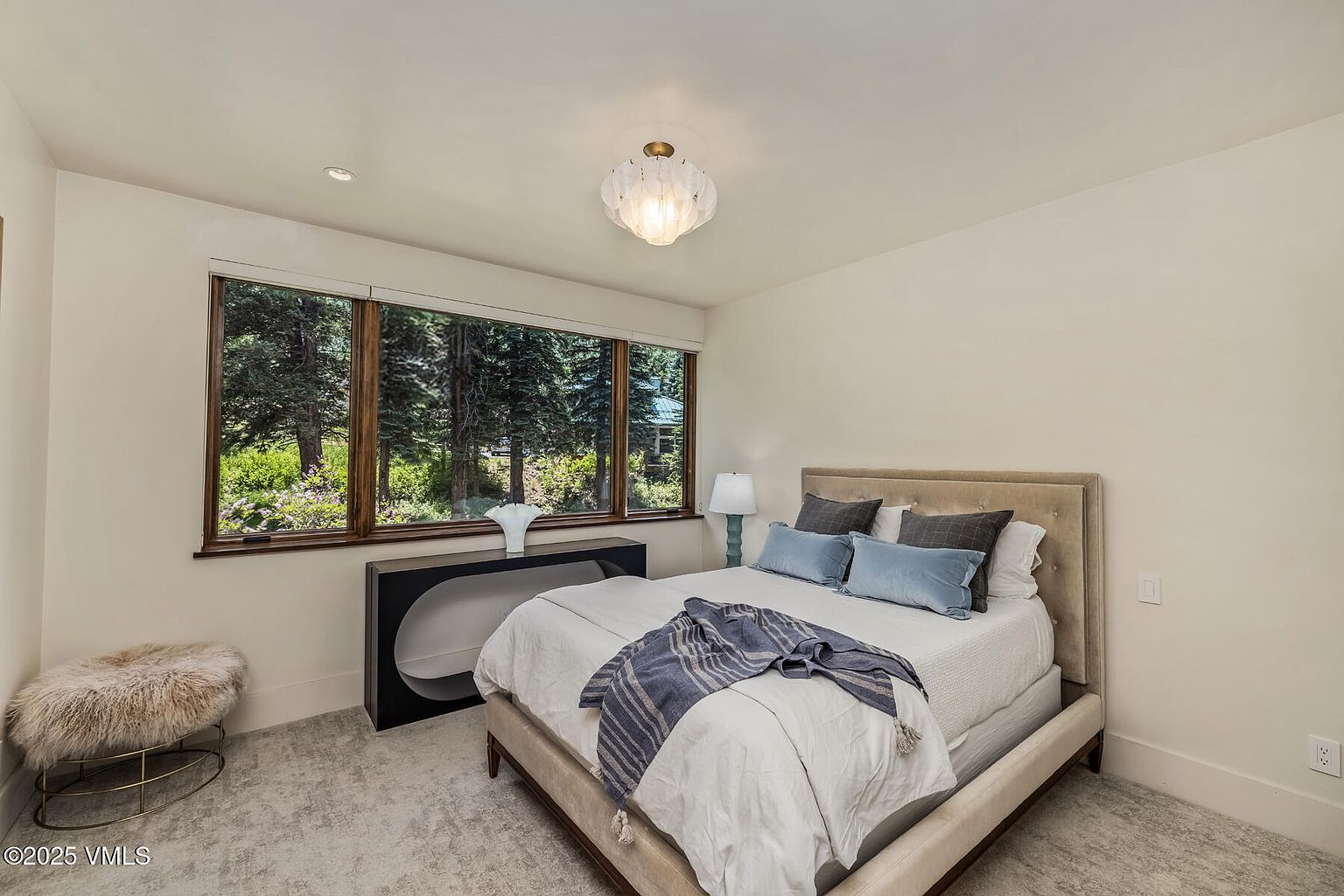 This serene bedroom features a neutral color palette with a plush, upholstered bed frame and soft, layered bedding. A large window provides a view of the surrounding forest, while a sleek, dark console table sits beneath it, topped with a decorative vase. The room is completed by a textured, circular ottoman and a unique, multi-faceted glass light fixture, creating a calm and inviting atmosphere.