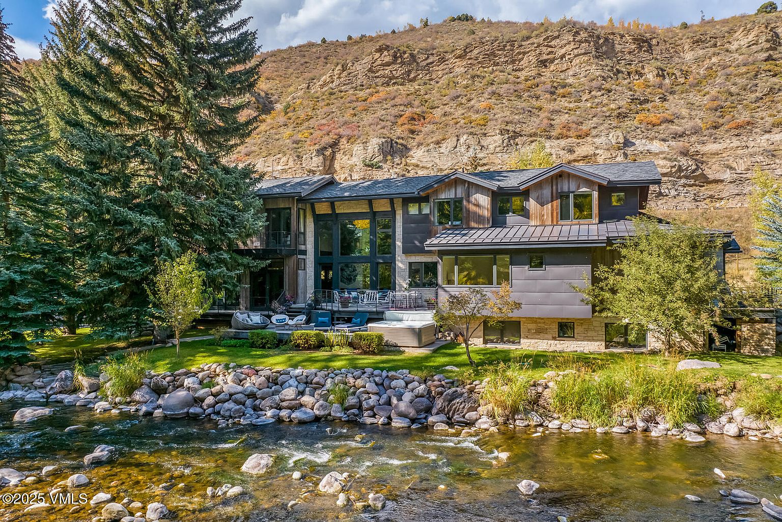 This stunning mountain residence is situated directly along a rushing river, featuring a multi-level architectural design that blends stone and dark metal siding. The rear elevation showcases a spacious deck with outdoor seating and a hot tub, all set against a dramatic, rugged cliffside backdrop. Large windows and glass doors create a seamless connection between the interior living spaces and the natural landscape.