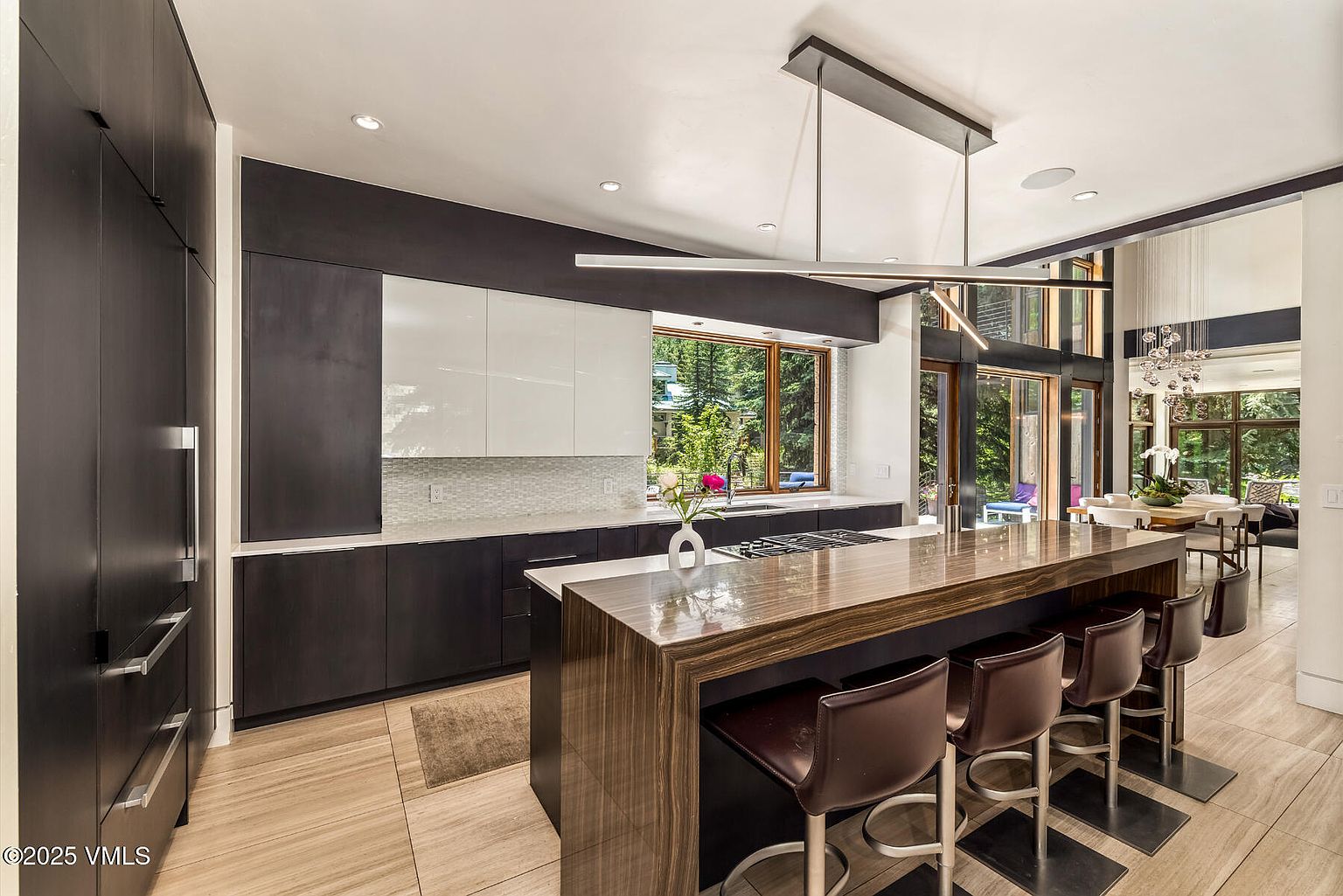 This modern kitchen features a striking contrast between dark, matte cabinetry and a sleek, wood-toned waterfall island. A long, linear pendant light fixture hangs above the island, which is paired with four leather-upholstered bar stools. Large windows provide a view of the lush greenery outside, while the open-concept layout flows seamlessly into an adjacent dining area.