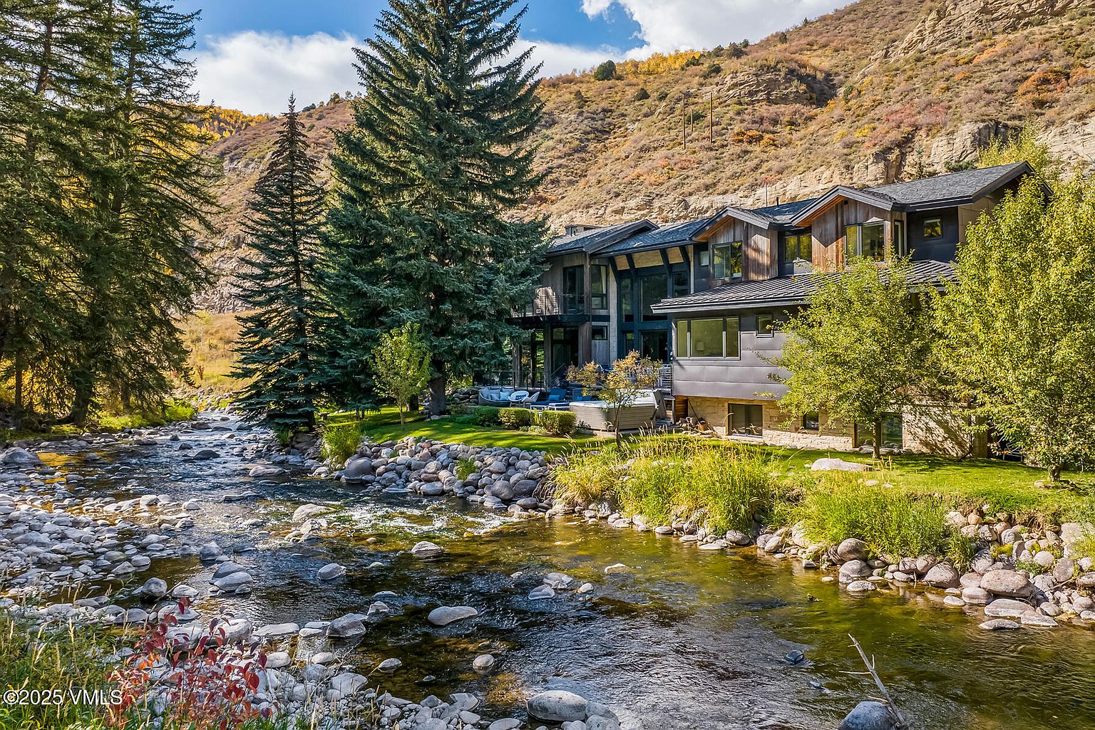 This stunning mountain residence is nestled directly along a scenic, rocky stream, offering a tranquil and private setting. The home features a contemporary architectural design with a mix of dark siding, natural wood accents, and large windows that capture the surrounding forest and hillside views. A landscaped lawn leads from the water's edge to a patio area equipped with a spa, creating a seamless connection between the luxury home and the natural landscape.