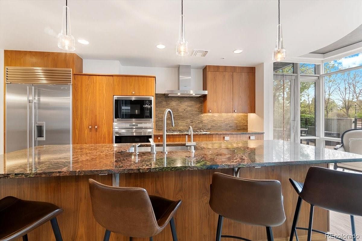 This is a modern kitchen featuring wood cabinetry, stainless steel appliances, and a large island with a unique countertop. Pendant lighting hangs above the island, and a large window provides natural light. The kitchen has a sleek and contemporary design.