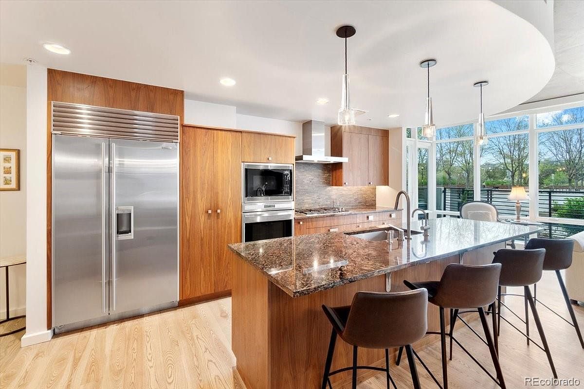 This is a well-lit, modern kitchen featuring wood cabinetry and stainless steel appliances, including a large refrigerator. A granite countertop island with bar seating is the focal point, illuminated by pendant lights. Large windows provide natural light and a view of the outdoors, creating a bright and inviting space.