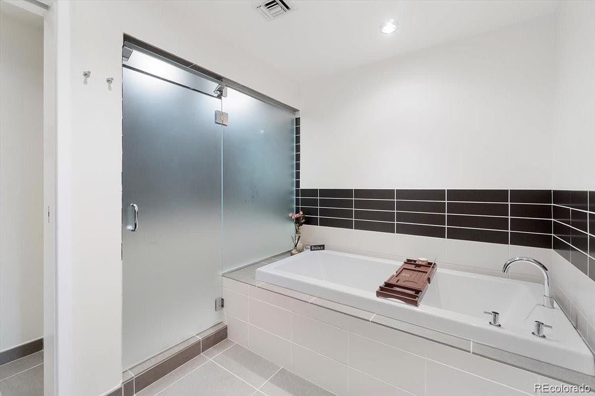 This is a primary bathroom featuring a modern bathtub with a brown wooden tray, a sleek chrome faucet, and a frosted glass shower enclosure. The walls are adorned with a combination of white and black horizontal tiles, creating a contemporary aesthetic. The overall impression is clean, bright, and relaxing.