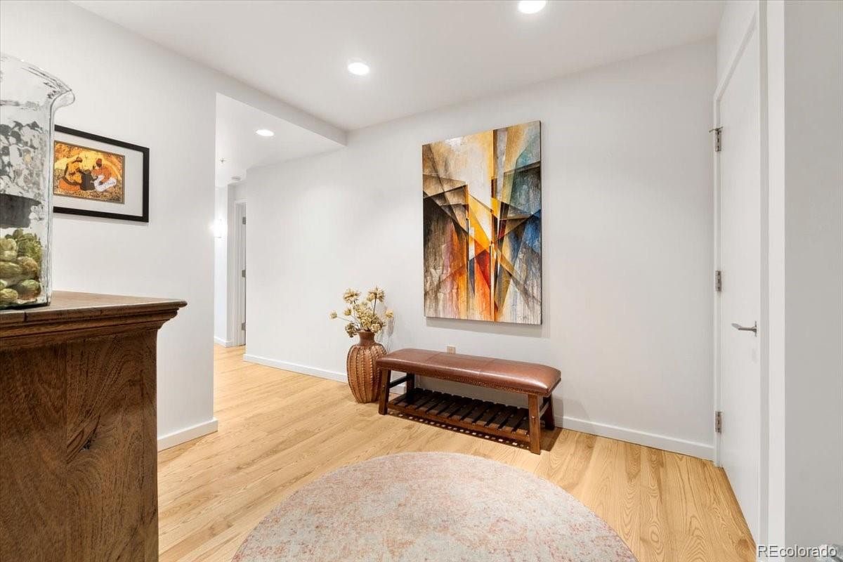 This is an interior shot of a hallway featuring light hardwood floors and white walls. An abstract painting hangs on the wall above a wooden bench with a leather cushion. A round rug sits on the floor, and a framed picture is visible on the left wall, adding a touch of personality to the space.