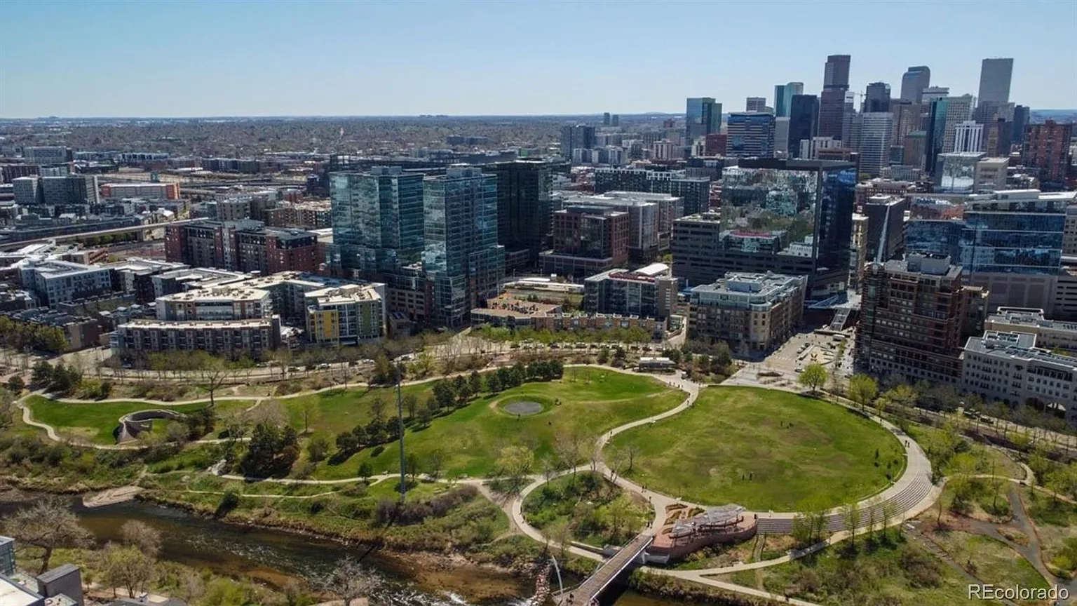 Aerial view showcasing a cityscape with a prominent green park. The urban landscape features modern buildings and skyscrapers in the background. A river flows alongside the park, adding to the scenic view.