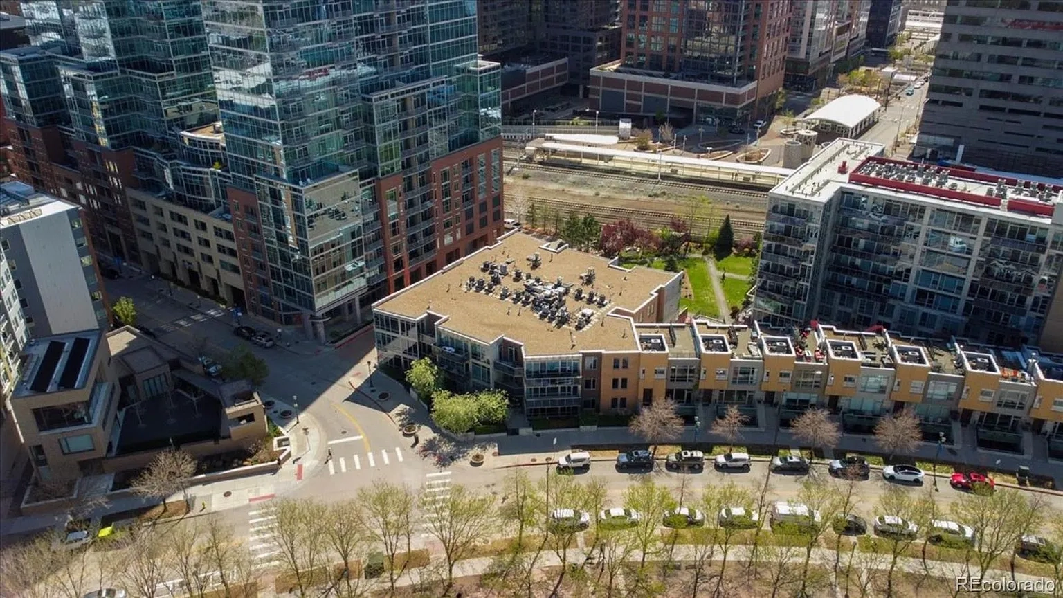 This aerial view showcases a dense urban environment with a mix of modern high-rise buildings and multi-story residential complexes. The architecture features lots of glass, brick, and some metal. The image also highlights streetscapes with rows of trees, providing a glimpse of the community surroundings.