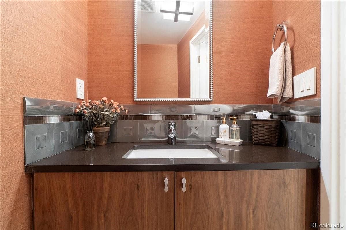 This is a well-lit bathroom featuring a wooden vanity with a dark countertop and a white sink. The walls are covered in a textured orange wallpaper, complemented by gray tiled backsplash. A mirror hangs above the sink, and a towel rack with towels is visible on the right side of the frame.