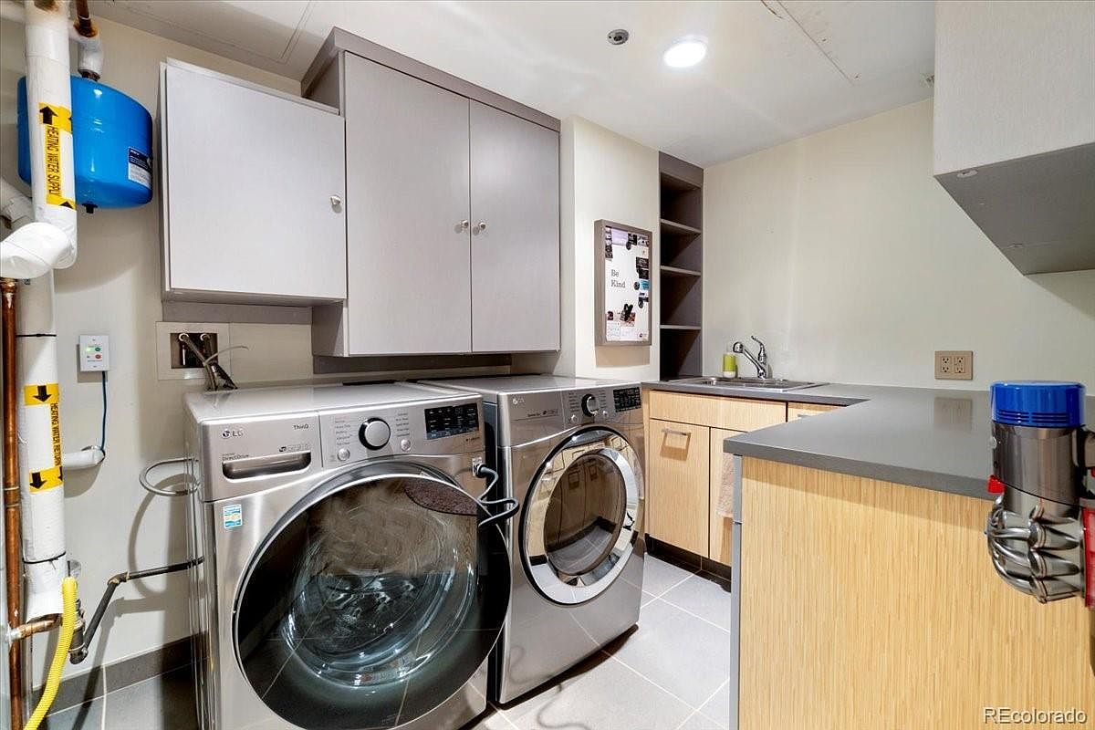 This is an interior shot of a laundry room featuring a front-loading washer and dryer set in a silver finish. Above the appliances are gray cabinets, and to the right, there's a sink with a countertop and additional cabinetry. The room is well-lit with a neutral color palette, creating a clean and functional space.