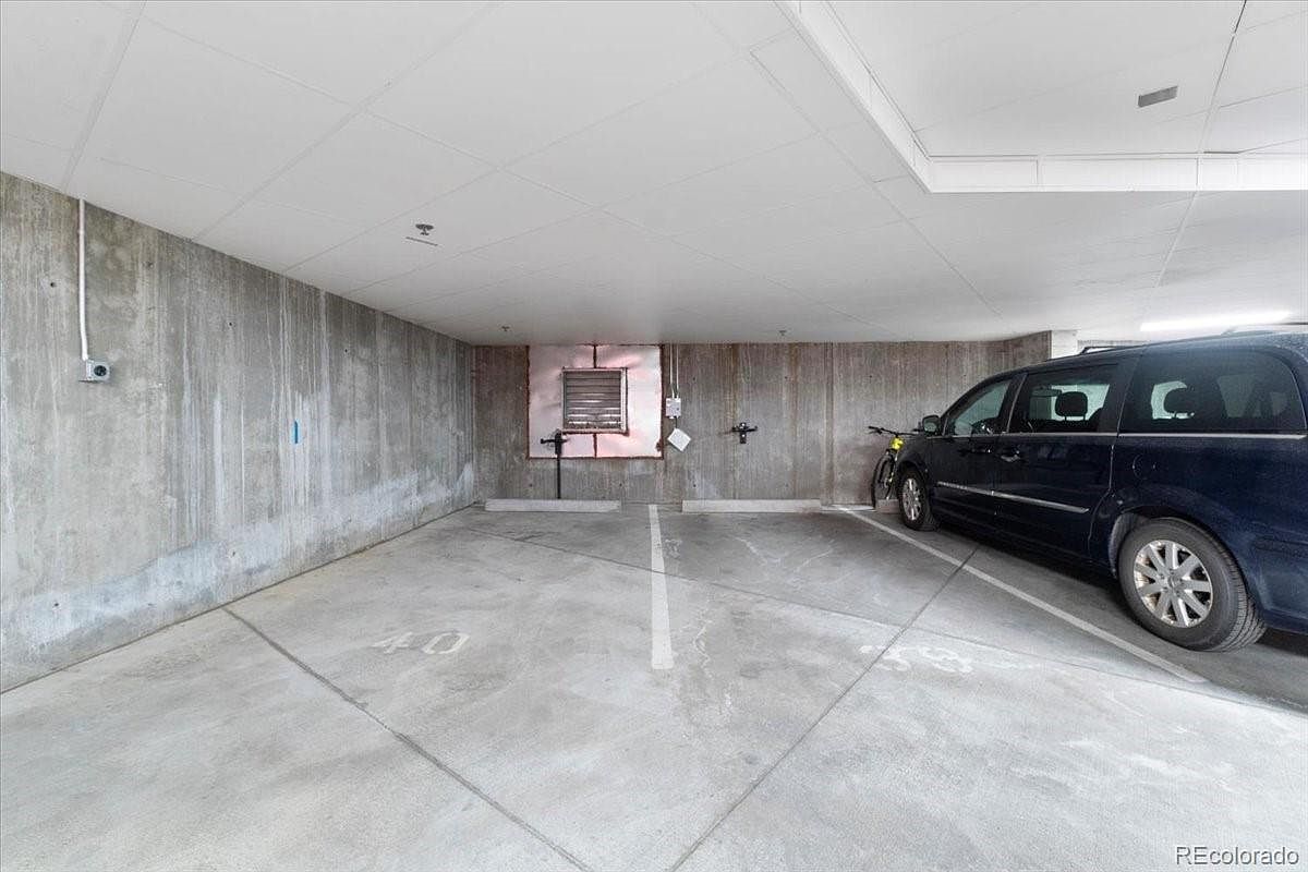 The image showcases an indoor parking garage space with concrete walls and flooring. A dark-colored minivan is parked on the right side, and a bicycle is leaning against the wall nearby. The space is well-lit, and parking lines are visible on the floor, indicating designated parking spots.
