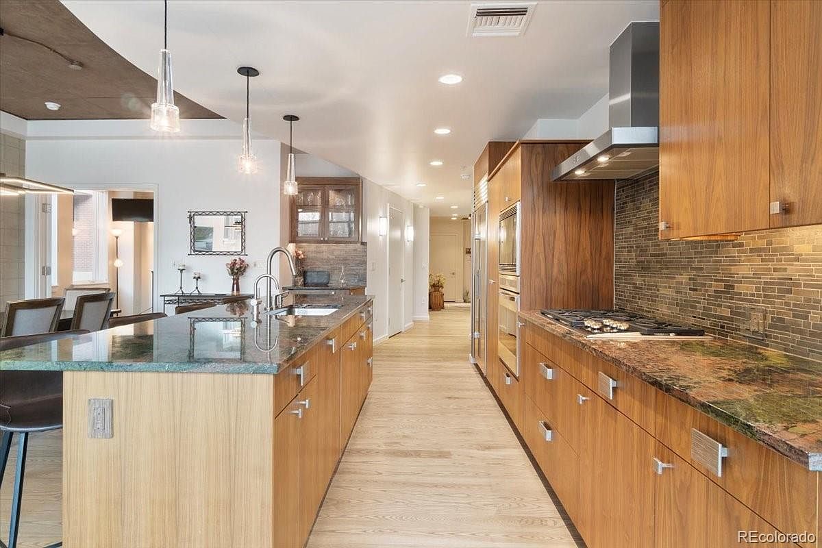 This is a well-lit, modern kitchen featuring wooden cabinetry and granite countertops. The kitchen island provides ample workspace and seating, while stainless steel appliances add a touch of elegance. The hardwood flooring and mosaic backsplash contribute to the kitchen's sophisticated aesthetic.