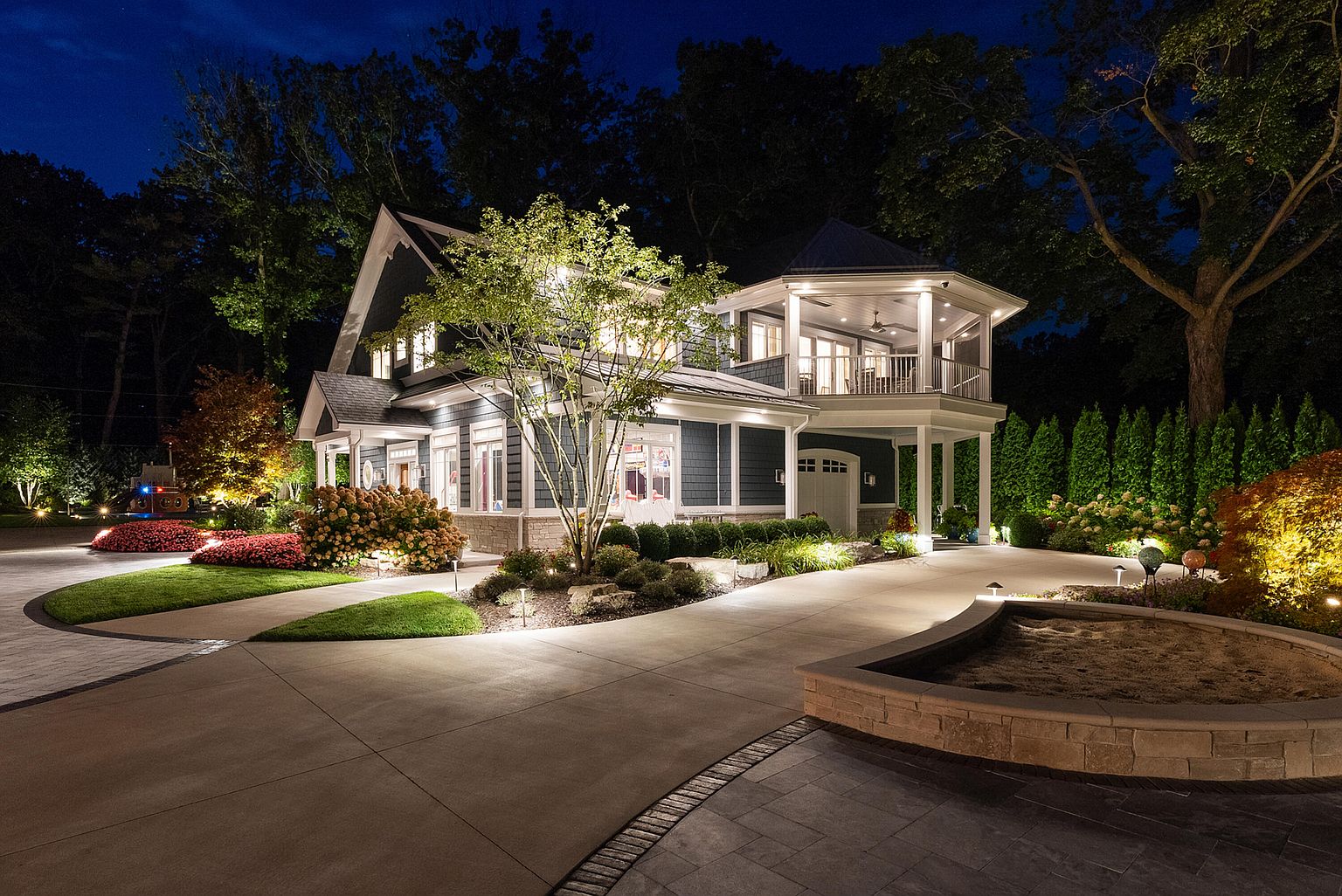 This is a beautifully illuminated front view of a two-story home at night. The house features a combination of siding and stone accents, with well-manicured landscaping and strategically placed lighting that highlights the architectural details. A circular driveway leads to the house, enhancing its curb appeal and creating a welcoming atmosphere.