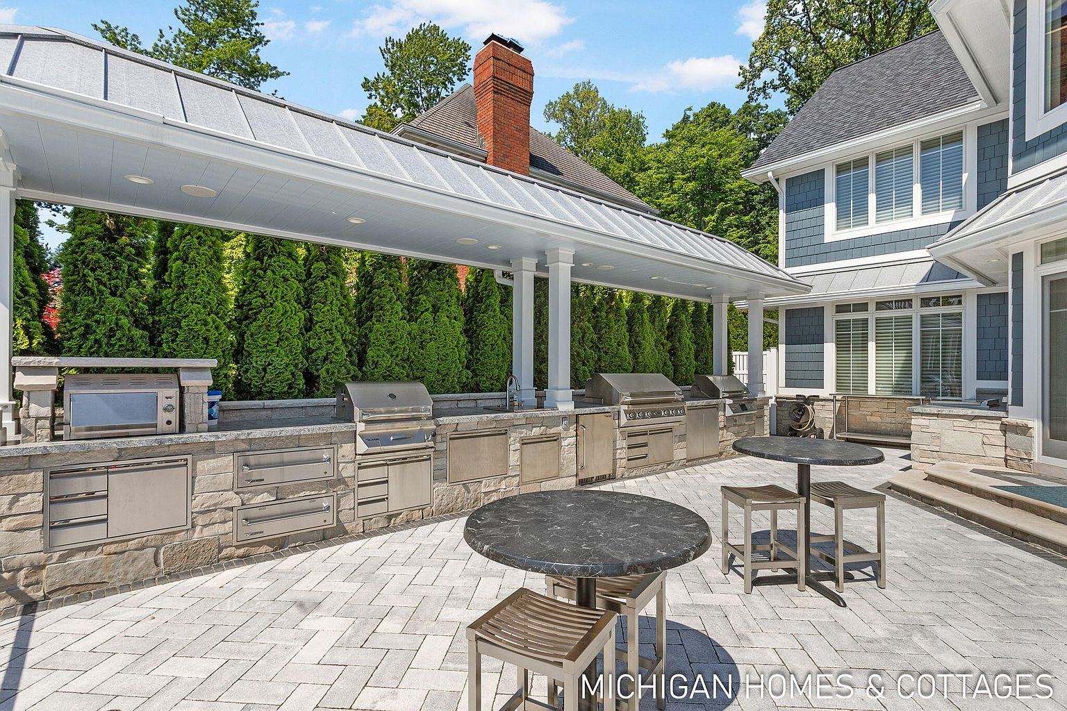 This image showcases a luxurious outdoor kitchen and patio area. The kitchen features stainless steel appliances built into a stone counter, covered by a stylish roof structure. The patio is paved with interlocking stones and includes outdoor dining tables, creating an inviting space for entertaining.