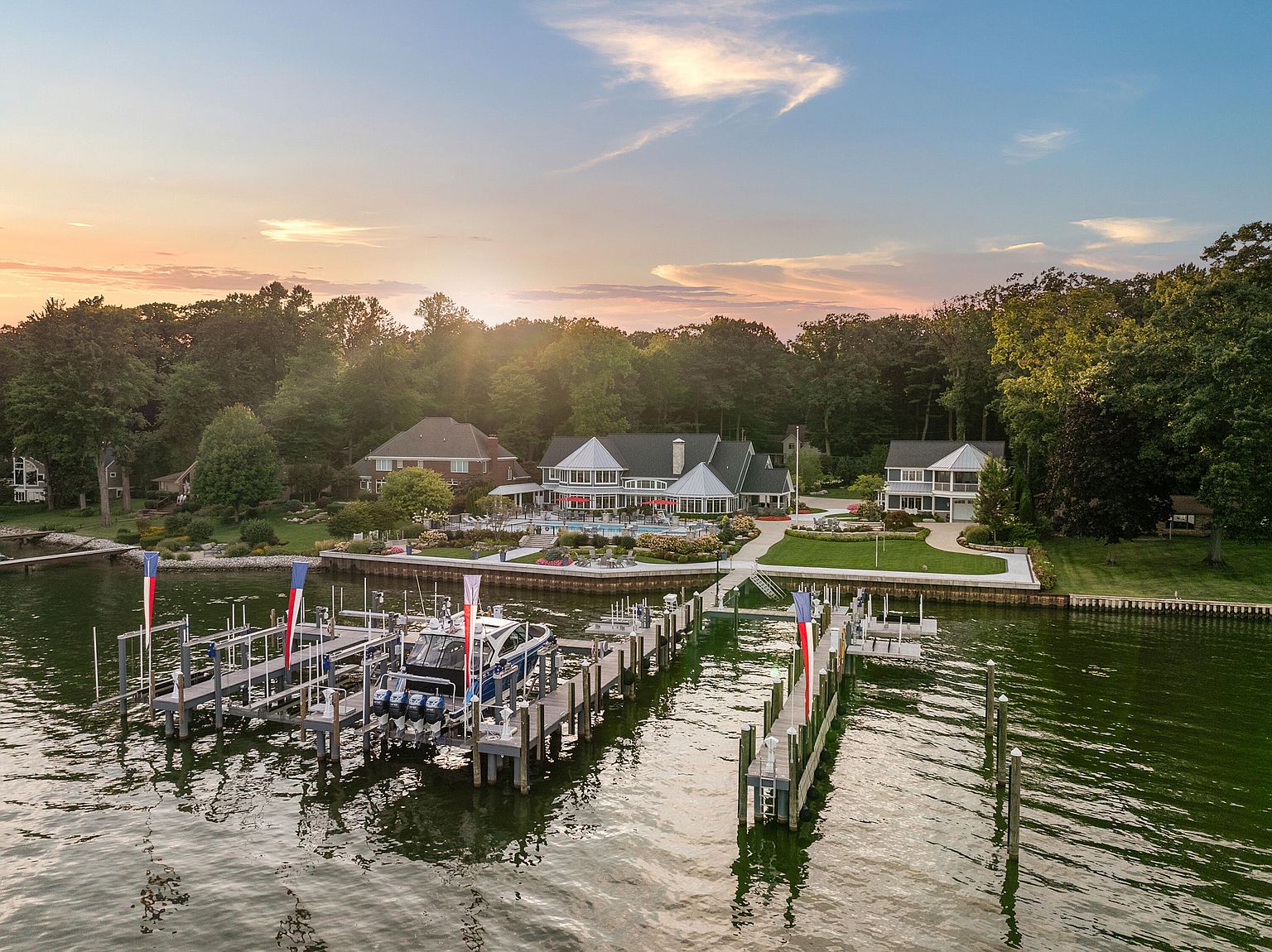 This aerial shot showcases a luxurious waterfront property at sunset. The home features a sprawling lawn, a pool area, and a private dock with a boat. The surrounding trees provide privacy, and the overall impression is one of opulence and tranquility.
