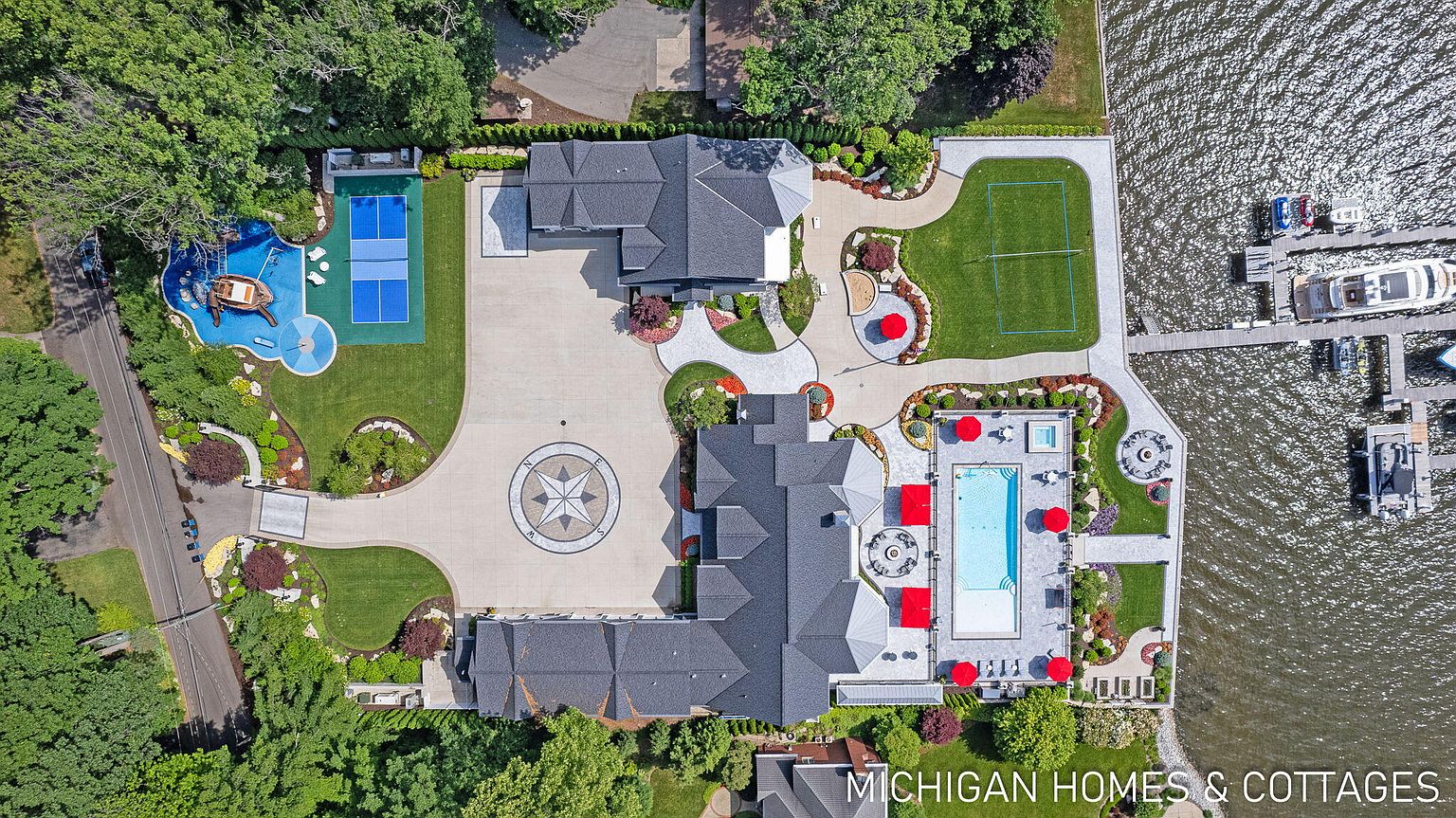 This aerial view showcases a luxurious waterfront estate with meticulously landscaped grounds. The property features a large main house, a swimming pool with red umbrellas, a tennis court, a playground, and a private dock with multiple boats. The circular driveway with a compass design adds a unique touch to the property's grand entrance.