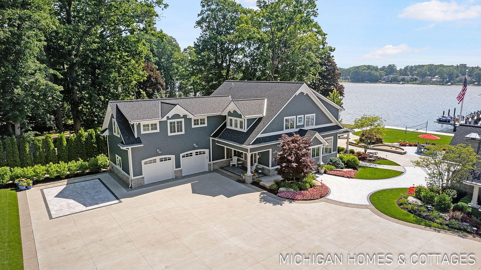 This aerial view showcases a beautifully landscaped property featuring a gray shingle-style home with white trim, a spacious driveway, and lush greenery. The house is situated near a lake, adding to the property's appeal. The overall impression is one of luxury and tranquility, highlighting the home's prime location and well-maintained exterior.