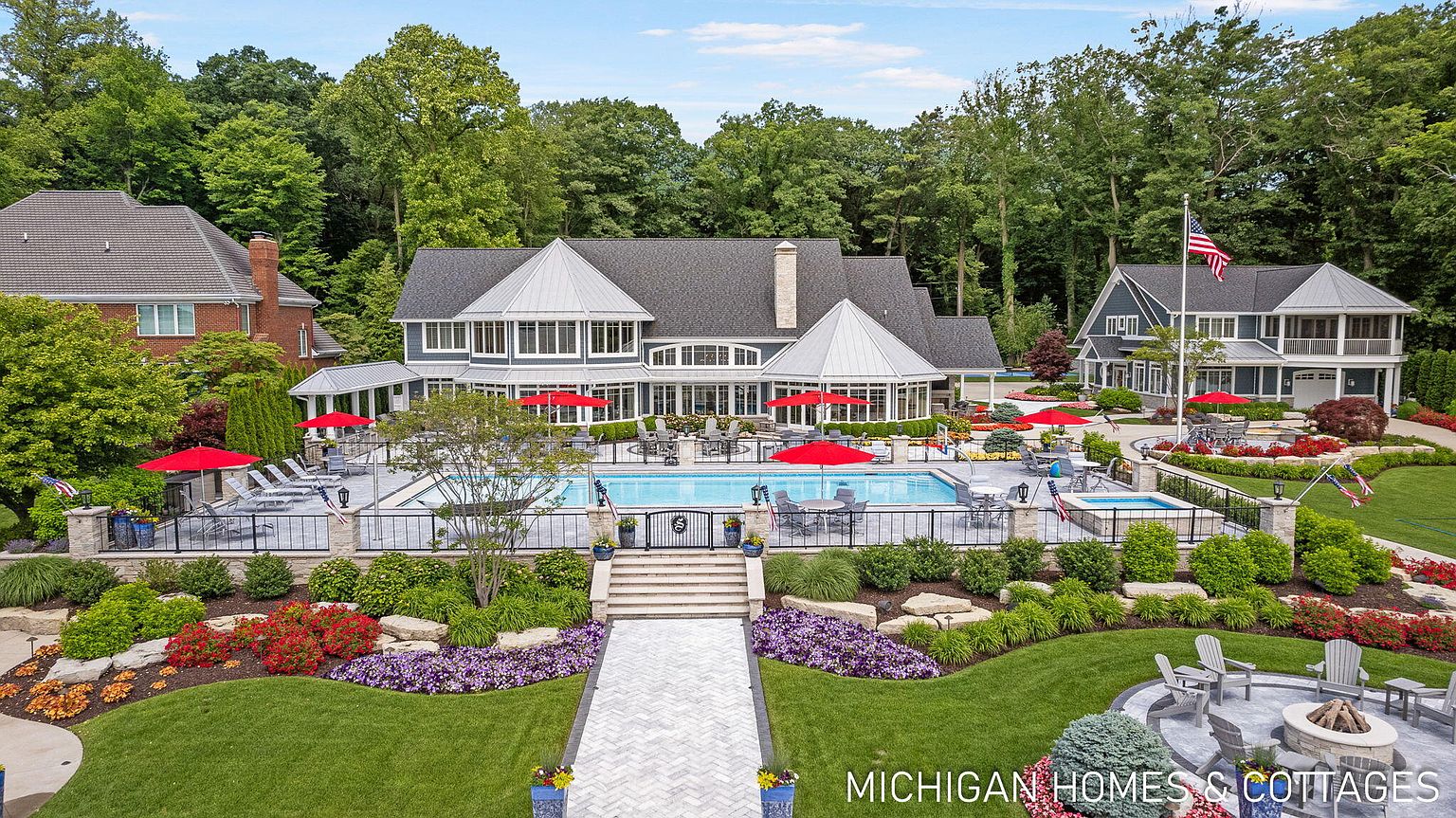 This is a stunning rear view of a luxurious home, showcasing a beautiful pool area with red umbrellas and lounge chairs, surrounded by meticulously landscaped gardens. The architecture features a combination of gray siding and white trim, with large windows offering ample natural light. A paved walkway leads to a fire pit area, enhancing the outdoor living space.