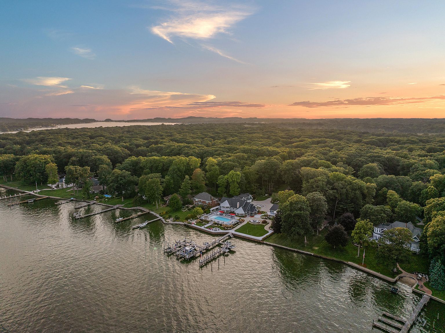 This aerial view showcases a luxurious waterfront property with a large house, a swimming pool, and multiple docks extending into the water. The property is surrounded by lush, dense forest, creating a sense of privacy and seclusion. The image captures a serene sunset, enhancing the overall appeal and highlighting the property's prime location and upscale features.