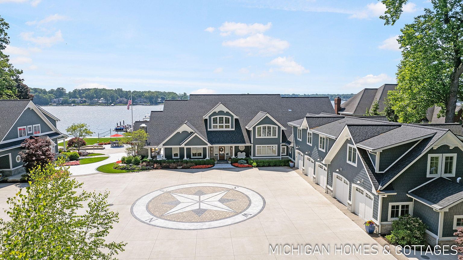 This aerial view showcases a grand waterfront estate with a circular driveway featuring a compass design. The home boasts a dark gray roof, gray siding, and multiple gabled dormers, exuding a luxurious and stately presence. Lush landscaping and a glimpse of the lake enhance the property's appeal.