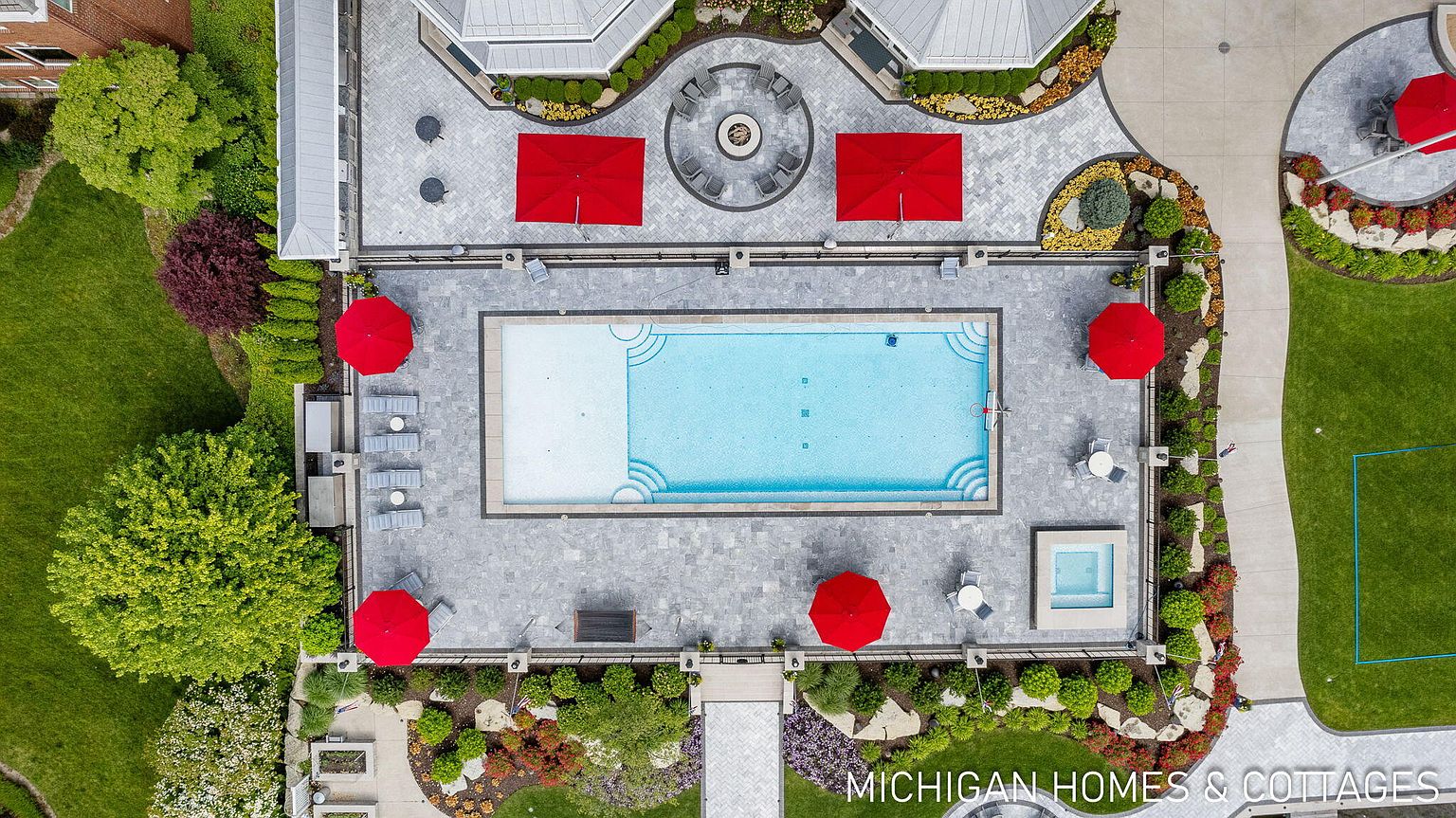 This aerial shot showcases a luxurious pool and spa area, complete with red umbrellas, lounge chairs, and meticulously landscaped surroundings. The pool features a light blue interior, while the spa is a smaller, square-shaped addition. The surrounding patio is paved with gray stone, and the landscaping includes a variety of plants, flowers, and decorative rocks, creating a resort-like atmosphere.