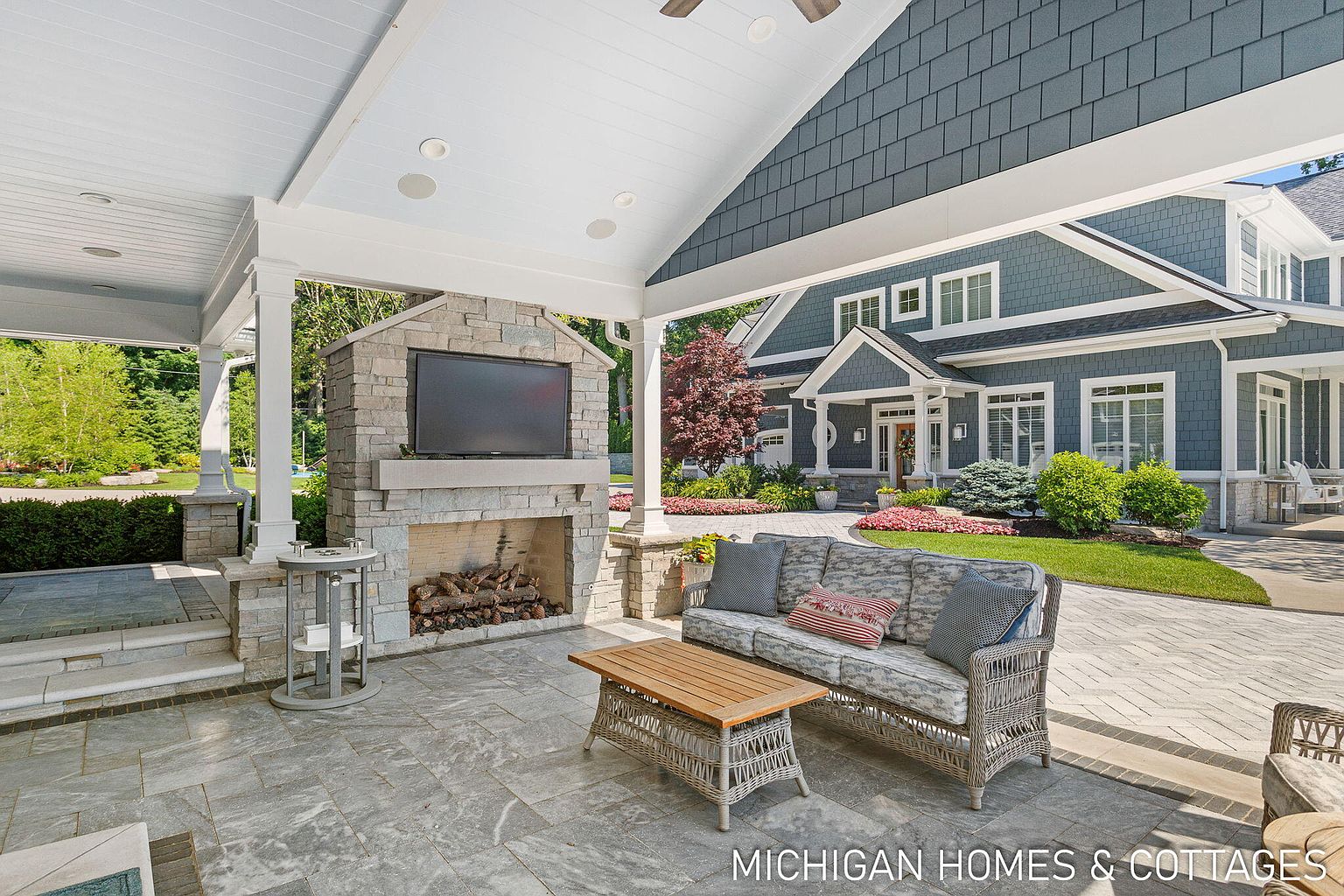 This image showcases a luxurious outdoor patio area, featuring a stone fireplace with a mounted television, comfortable wicker seating, and a wooden coffee table. The patio is covered by a white-painted roof supported by elegant columns, offering shelter and style. The well-manicured lawn and the house's exterior, with its blue siding and white trim, create an inviting and upscale outdoor living space.