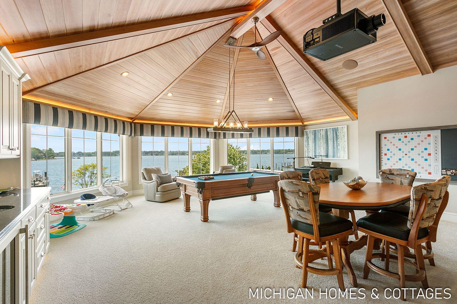 This interior shot showcases a spacious living room with a unique architectural design, featuring a high, wood-paneled ceiling and large windows offering a waterfront view. The room includes a pool table, a dining area with a round table and chairs, and a projector mounted on the ceiling, creating a luxurious and entertainment-focused space. The overall impression is one of elegance and comfort, perfect for relaxation and social gatherings.