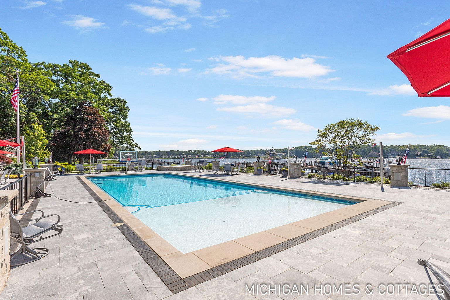 This image showcases a luxurious outdoor pool area, complete with a rectangular pool featuring a light blue and white tile design. The pool is surrounded by a spacious patio made of gray stone tiles, and red umbrellas provide shade. In the background, there's a view of a lake or river with boats docked, enhancing the property's appeal for waterfront living.