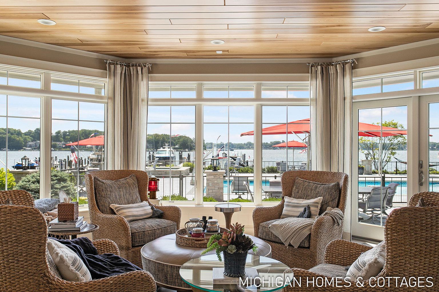 This is an interior shot of a sunroom or living room with a waterfront view. The room is furnished with wicker chairs and a round ottoman-style coffee table. Large windows offer a panoramic view of the water, a dock, and a swimming pool, creating a bright and inviting atmosphere.