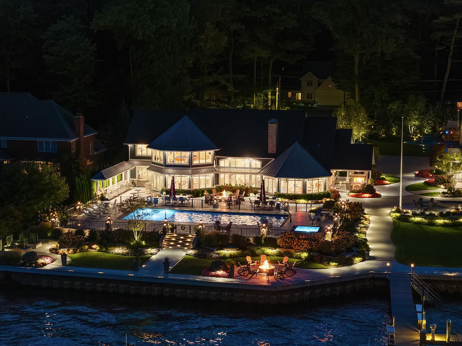 This aerial view showcases a luxurious waterfront property at night, featuring a large house with illuminated windows, a swimming pool, a spa, and a fire pit area. The landscape is meticulously designed with manicured gardens and pathways, creating an inviting outdoor living space. A private dock extends into the water, enhancing the property's appeal for waterfront living.