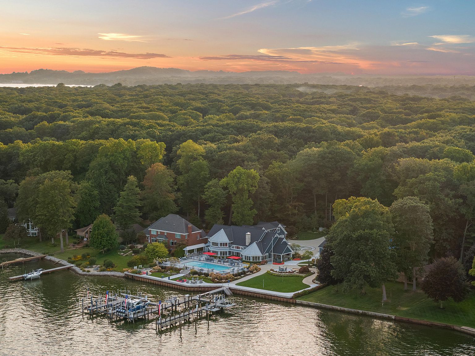 This aerial view showcases a luxurious waterfront property at sunset. The estate features a large house with a pool, manicured lawns, and a private dock with multiple boats. The property is surrounded by dense forest, offering privacy and a serene setting.