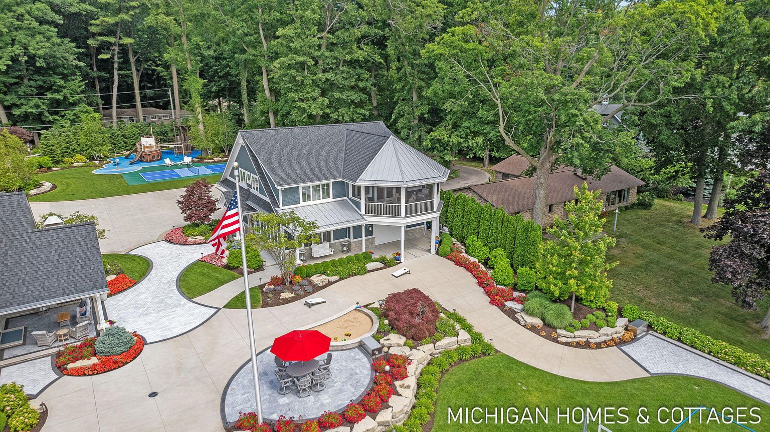 This aerial shot showcases a beautifully landscaped property featuring a two-story house with a screened porch, a separate guest house, and a playground area. The meticulously maintained grounds include winding walkways, vibrant flower beds, mature trees, and an American flag, creating a luxurious and inviting outdoor space. A red umbrella covers a table and chairs on a patio.