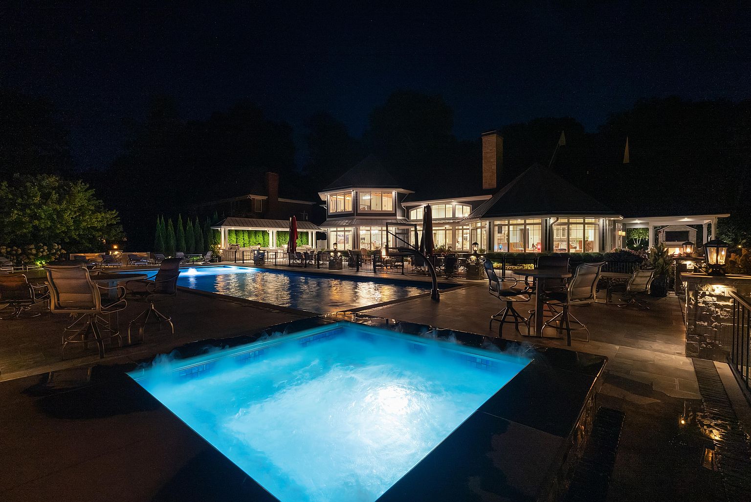 This image showcases a luxurious backyard at night, featuring a well-lit pool and spa area. The pool and spa are illuminated with a vibrant blue light, creating a serene and inviting atmosphere. The surrounding patio is furnished with outdoor seating, and the house in the background has large windows that allow light to spill out, enhancing the overall ambiance.
