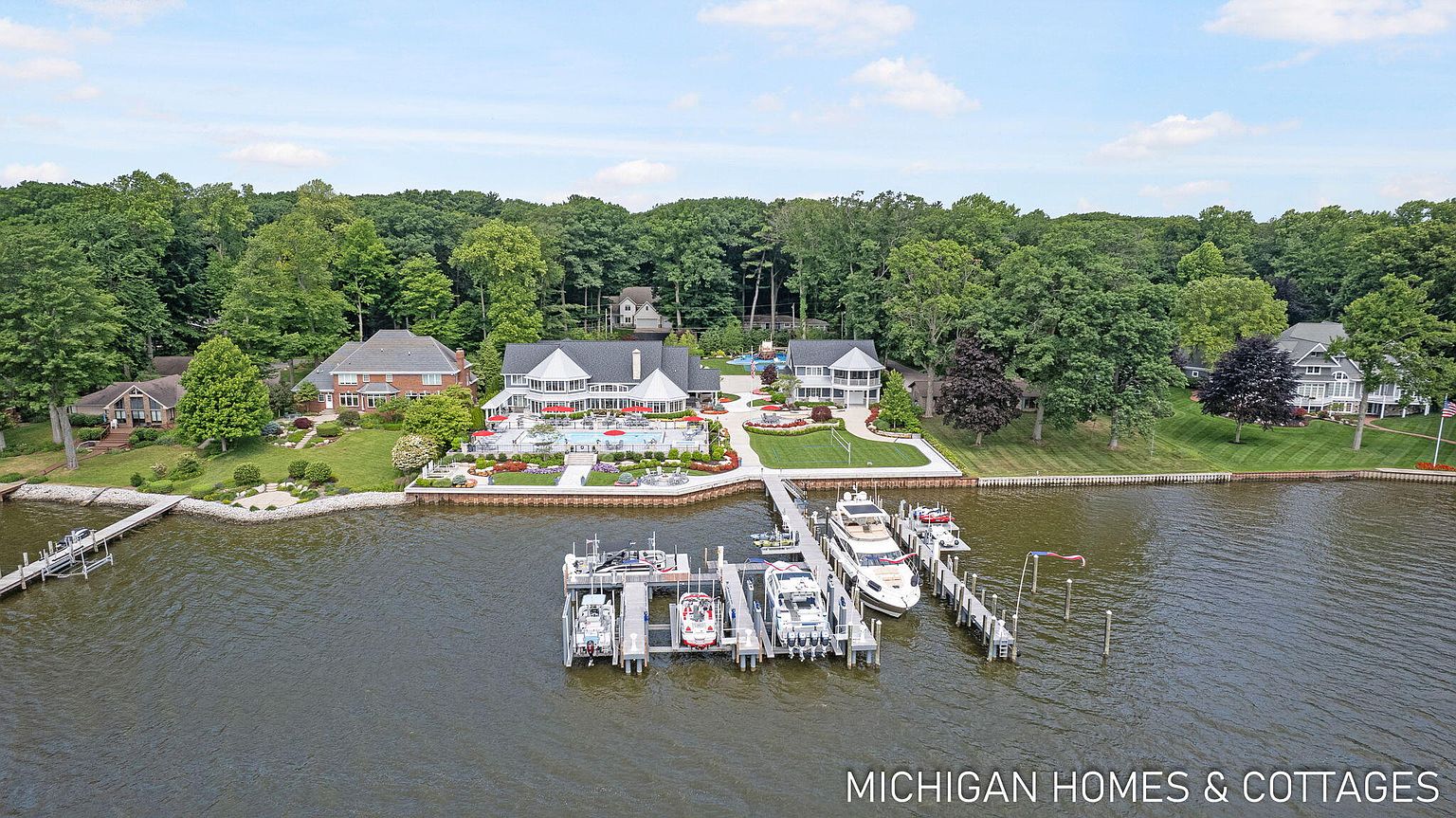 This aerial view showcases a luxurious waterfront property with multiple buildings, a swimming pool, meticulously landscaped gardens, and a private dock with several boats. The main house features a gray roof and white exterior, complemented by lush greenery and mature trees surrounding the estate. The overall impression is one of opulence and serene lakeside living.