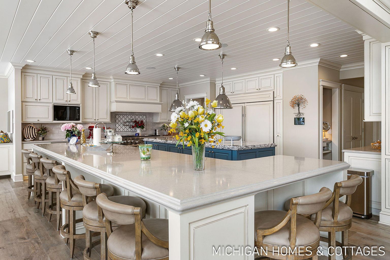 This is a bright and spacious kitchen featuring a large island with seating, white cabinetry, and stainless steel appliances. The island has a light-colored countertop and a contrasting blue base, adding a pop of color. Pendant lights hang above the island, illuminating the space, and the overall design is elegant and inviting.