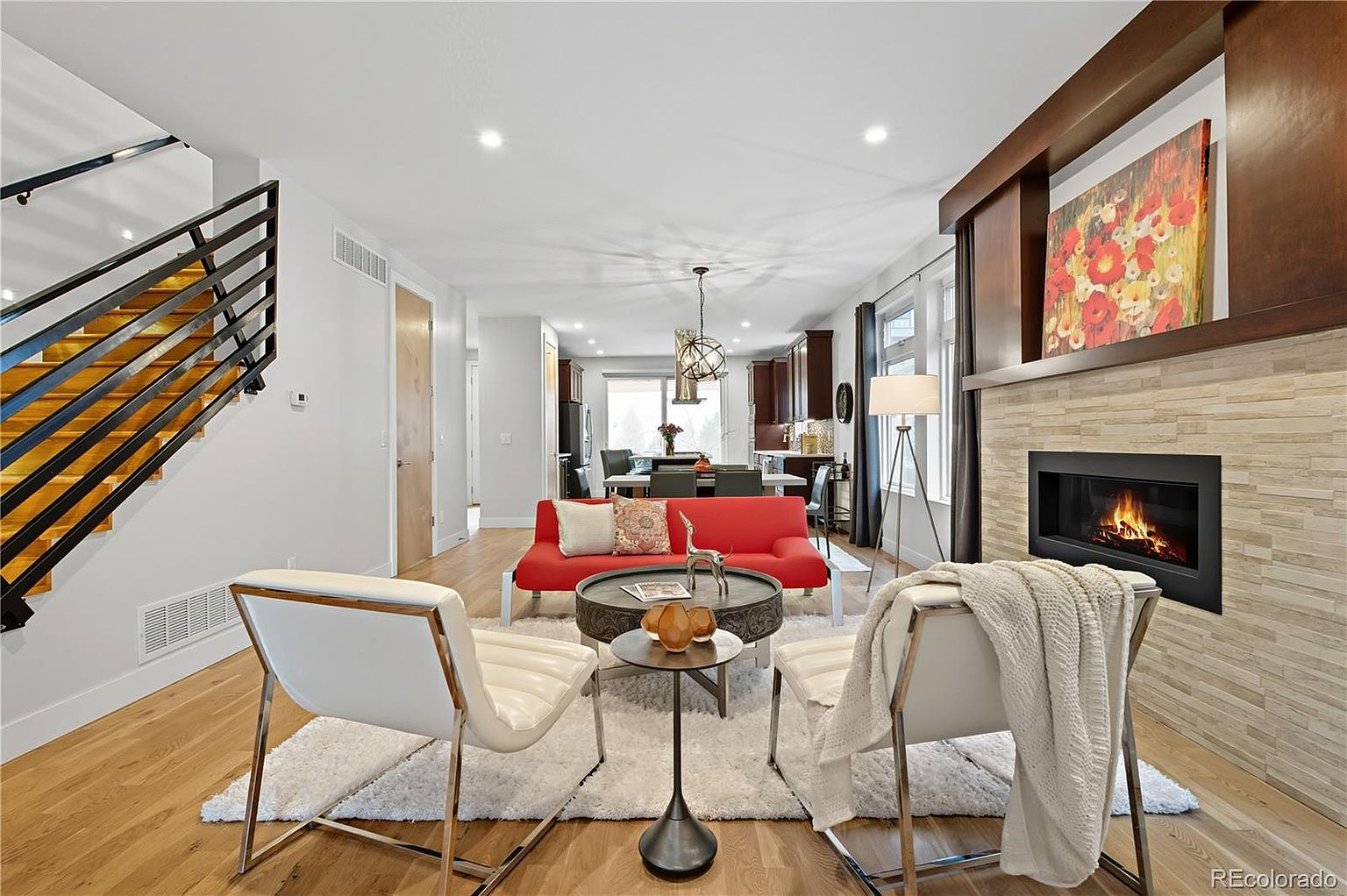 This is an interior shot of a modern living room featuring a red sofa, two white chairs, and a fireplace with a stone surround. The room has hardwood floors and a neutral color palette, with a staircase visible on the left. The overall impression is stylish and inviting, perfect for a cozy and contemporary home.