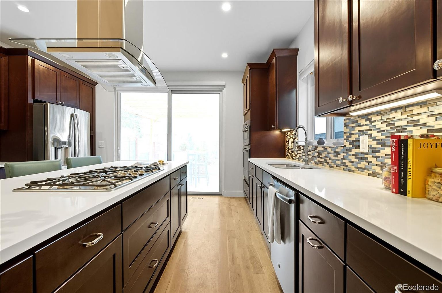 This is a well-lit kitchen featuring dark wood cabinetry, white countertops, and stainless steel appliances. The kitchen island has a built-in gas cooktop. A sliding glass door provides natural light and access to the outside, while a mosaic backsplash adds a touch of color and texture.